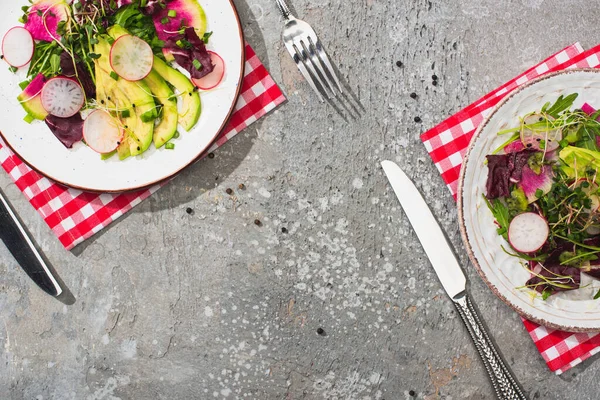 Vue de dessus de salade de radis frais avec légumes verts et avocat servi sur des assiettes sur la surface de béton gris avec couverts et serviettes — Photo de stock