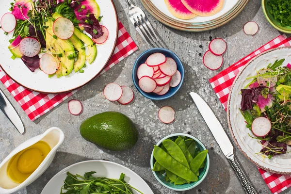 Vue de dessus de salade de radis frais avec légumes verts et avocat servi sur des assiettes sur la surface de béton gris avec des ingrédients, couverts et serviettes — Photo de stock