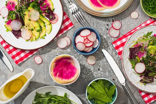 Vue de dessus de salade de radis frais avec légumes verts et avocat servi sur des assiettes sur la surface de béton gris avec des ingrédients, couverts et serviettes — Photo de stock