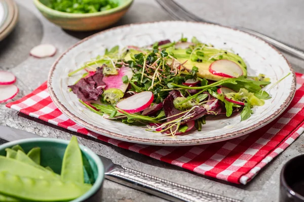 Salada de rabanete fresco com verduras e abacate servido em guardanapo com talheres perto de ingredientes em tigelas na superfície de concreto cinza — Fotografia de Stock