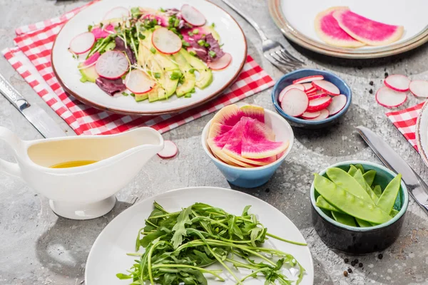 Fresh radish salad with greens and avocado in plate served on napkin with cutlery near ingredients in bowls on grey concrete surface — Stock Photo