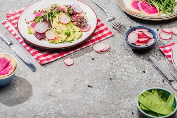 Salada de rabanete fresco com verduras e abacate servido em guardanapo com talheres perto de ingredientes em tigelas na superfície de concreto cinza — Fotografia de Stock