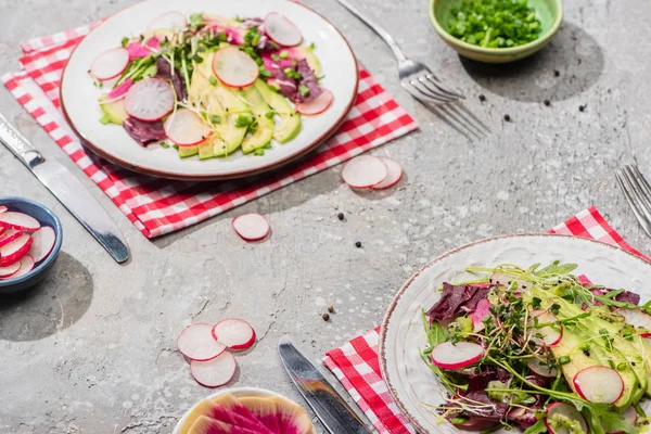 Insalata di ravanello fresco con verdure e avocado in piatti serviti su tovaglioli con posate vicino ingredienti in ciotole su superficie di cemento grigio — Foto stock