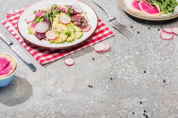 Fresh radish salad with greens and avocado served on napkin with cutlery on grey concrete surface — Stock Photo