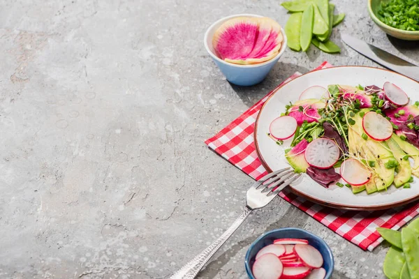 Salada de rabanete fresco com verduras e abacate servido em guardanapo com talheres perto de ingredientes em tigelas na superfície de concreto cinza — Fotografia de Stock