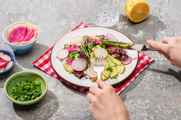 Vista cortada de mulher comendo salada de rabanete fresco com verduras e abacate em guardanapo na superfície de concreto cinza com ingredientes em tigelas — Fotografia de Stock