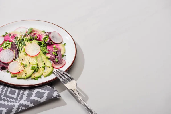 Свежий салат из редиса с зеленью и авокадо на серой поверхности с салфеткой и вилкой — стоковое фото
