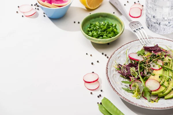 Fresh radish salad with greens and avocado on white surface with ingredients in bowls, water and fork — Stock Photo