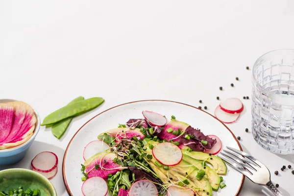 Свежий салат из редиса с зеленью и авокадо на белой поверхности с ингредиентами в мисках, воде и вилке — стоковое фото