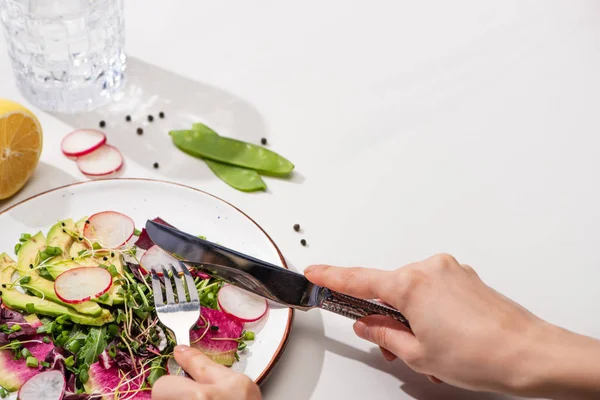 Cropped view of woman eating fresh radish salad with greens and avocado on white surface with water — Stock Photo