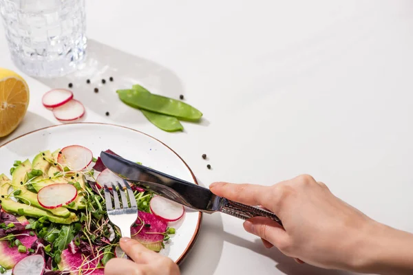 Vista recortada de la mujer comiendo ensalada de rábano fresco con verduras y aguacate en la superficie blanca con agua - foto de stock