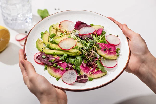 Vista ritagliata di donna che tiene l'insalata di ravanello fresca con verdi e avocado sul piatto — Foto stock