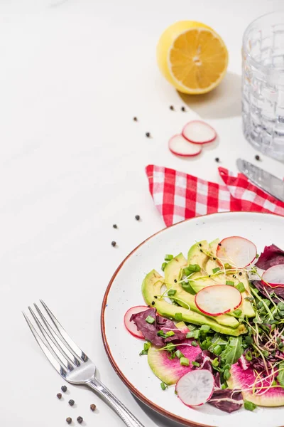 Селективний фокус свіжого салату з редькою з зеленню та авокадо на тарілці на білій поверхні з водою, лимоном та столовими приборами — стокове фото
