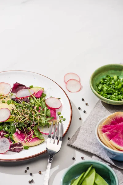 Frischer Rettichsalat mit Gemüse und Avocado auf Teller auf weißer Oberfläche mit Zutaten in Schüsseln und Gabel — Stockfoto