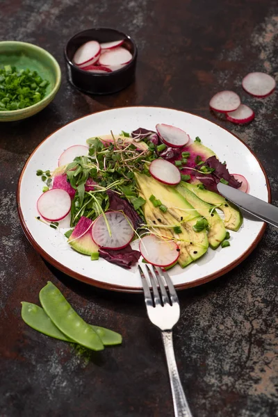 Focus selettivo di insalata di ravanello fresco con verdure e avocado su piatto servito su superficie intemperie con posate e ingredienti — Foto stock