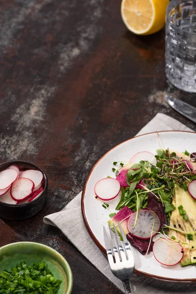 Fresh radish salad with greens and avocado on plate served on weathered surface with cutlery, lemon and water — Stock Photo