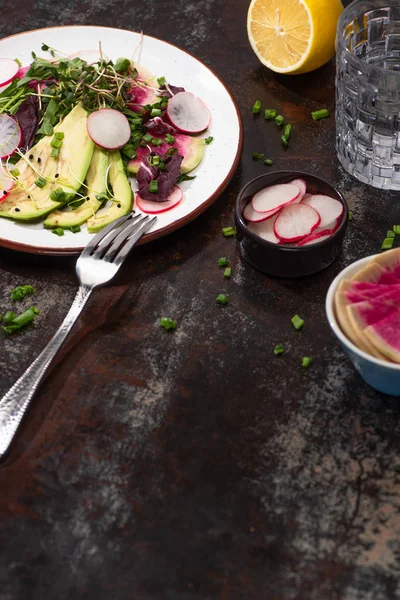 Свежий салат из редиса с зеленью и авокадо на тарелке на выветренной поверхности с вилкой, лимоном и водой — стоковое фото