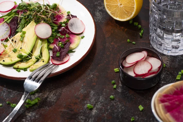 Insalata di ravanello fresco con verdure e avocado sul piatto su superficie intemperie con forchetta, limone e acqua — Foto stock