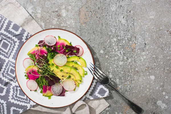Vista dall'alto di insalata di ravanello fresca con verdure e avocado vicino a forchetta su superficie di cemento grigia con tovaglioli — Foto stock