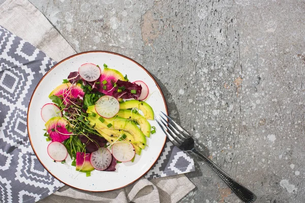 Vista superior de ensalada de rábano fresco con verduras y aguacate cerca de tenedor en superficie de hormigón gris con servilletas - foto de stock