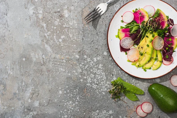 Top view of fresh radish salad with greens and avocado near fork and vegetables on grey concrete surface — Stock Photo