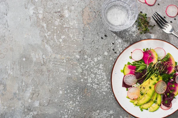 Top view of fresh radish salad with greens and avocado near fork and water on grey concrete surface — Stock Photo