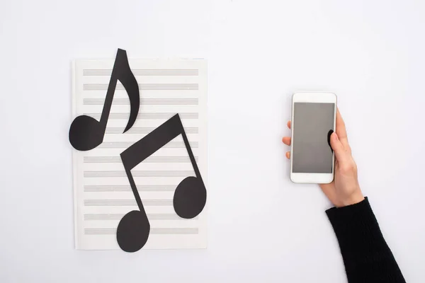 Cropped view of woman holding smartphone near paper cut notes on music book on white background — Stock Photo