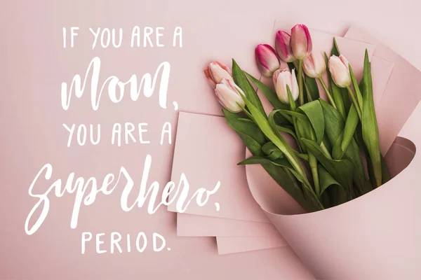 Vista superior de los tulipanes de primavera envueltos en papel sobre fondo rosa, si eres una madre, eres una ilustración de superhéroe - foto de stock