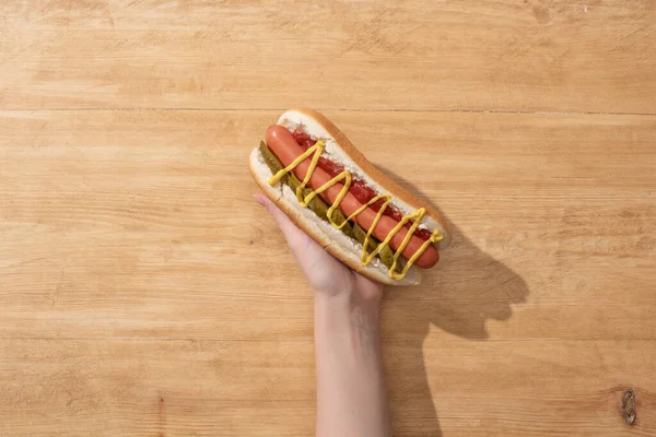 Abgeschnittene Ansicht einer Frau, die köstlichen Hot Dog mit Essiggurken, Senf und Ketchup auf einem Holztisch hält — Stockfoto