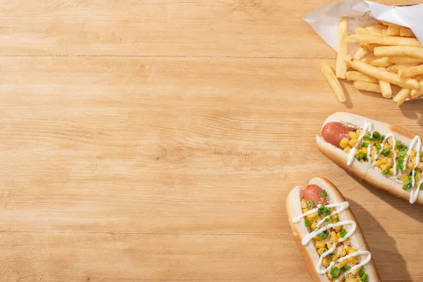 Top view of delicious hot dogs with corn, green onion and mayonnaise near french fries on wooden table — Stock Photo