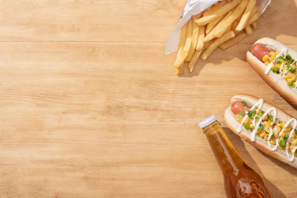 Vista superior de deliciosos cachorros-quentes com milho, cebola verde e maionese perto de batatas fritas e cerveja na mesa de madeira — Fotografia de Stock