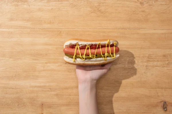 Partial view of woman holding delicious hot dog with pickles, mustard and ketchup on wooden table — Stock Photo