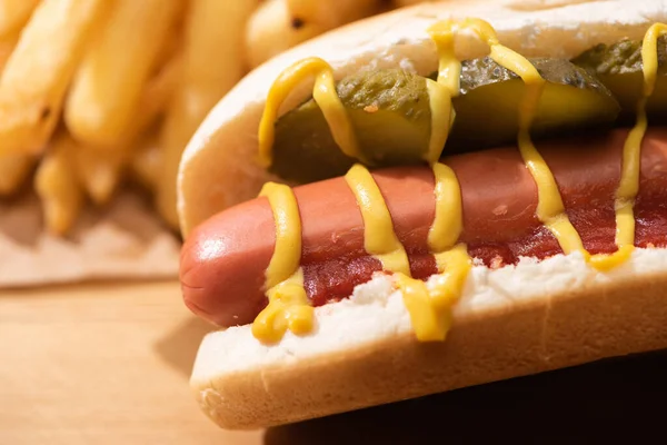 Vista de perto de delicioso cachorro-quente com picles, ketchup, mostarda perto de batatas fritas na mesa de madeira — Fotografia de Stock