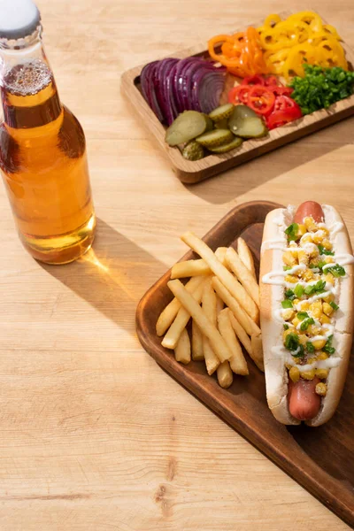 Delicious hot dog near board with sliced vegetables, beer and french fries on wooden table — Stock Photo