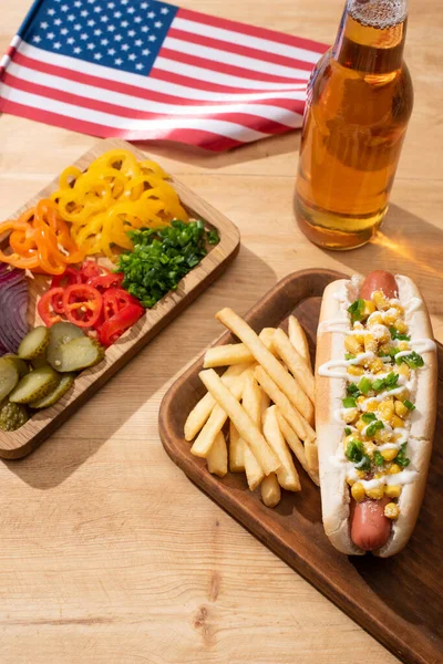 Delicious hot dog near american flag, board with sliced vegetables, beer and french fries on wooden table — Stock Photo