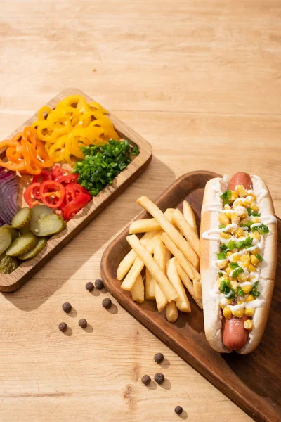 Delicious hot dog near board with sliced vegetable and french fries on wooden table — Stock Photo