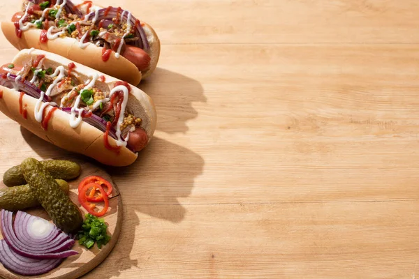 Deliciosos cachorros-quentes com cebola vermelha, bacon e mostarda Dijon perto do tabuleiro com legumes na mesa de madeira — Fotografia de Stock