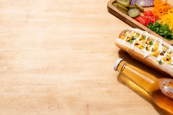 Delicious hot dog with corn, green onion and mayonnaise near bottle of beer and board with vegetables on wooden table — Stock Photo