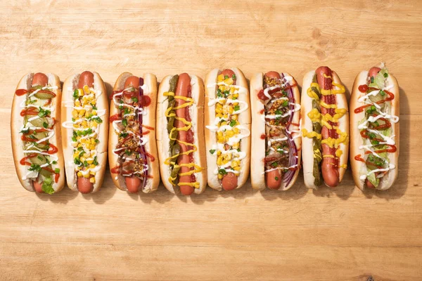 Blick von oben auf verschiedene leckere Hot Dogs mit Gemüse und Soßen auf Holztisch — Stockfoto