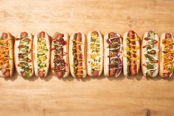 Blick von oben auf verschiedene leckere Hot Dogs mit Gemüse und Soßen auf Holztisch — Stockfoto