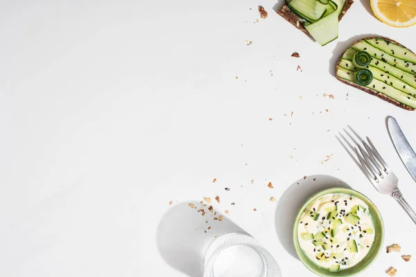 Vue de dessus des toasts au concombre frais près du citron, de l'eau, des couverts et du yaourt sur fond blanc — Photo de stock