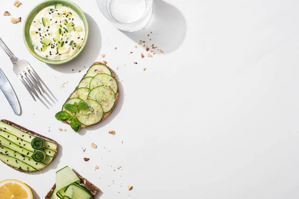 Vista dall'alto dei toast di cetrioli freschi vicino a limone, acqua, posate e yogurt su sfondo bianco — Foto stock
