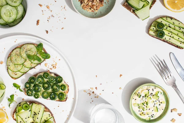 Vista superior de tostadas de pepino fresco en el plato cerca de limón, agua, cubiertos y yogur sobre fondo blanco - foto de stock