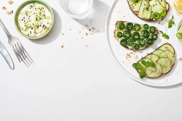 Vista superior de tostadas de pepino fresco en el plato cerca de limón, agua, cubiertos y yogur sobre fondo blanco - foto de stock