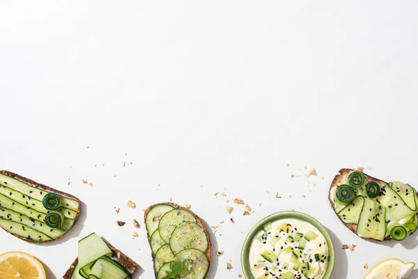 Top view of fresh cucumber toasts with seeds, mint and basil leaves and lemon near bowl of yogurt on white background — Stock Photo