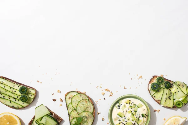 Vue de dessus des toasts au concombre frais avec des graines, des feuilles de menthe et de basilic et du citron près du bol de yaourt sur fond blanc — Photo de stock