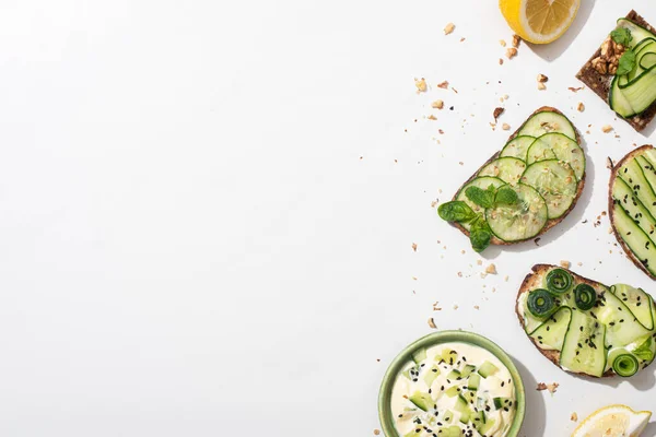 Top view of fresh cucumber toasts with seeds, mint and basil leaves and lemon near bowl of yogurt on white background — Stock Photo