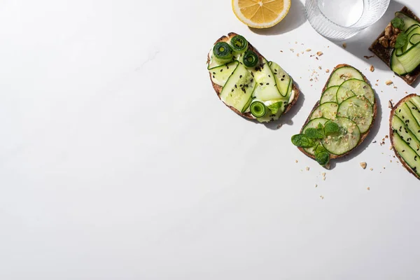 Top view of fresh cucumber toasts with seeds, mint and basil leaves, lemon near water on white background — Stock Photo