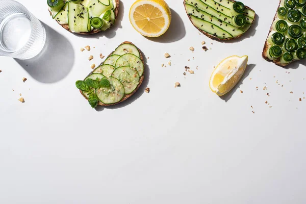 Vista dall'alto dei toast di cetrioli freschi con semi, menta e foglie di basilico, limone vicino all'acqua su sfondo bianco — Foto stock