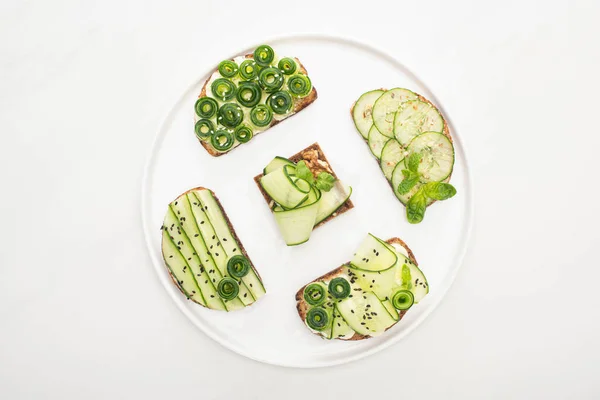 Vue de dessus des toasts au concombre frais avec des graines, de la menthe et des feuilles de basilic sur plaque ronde sur fond blanc — Photo de stock